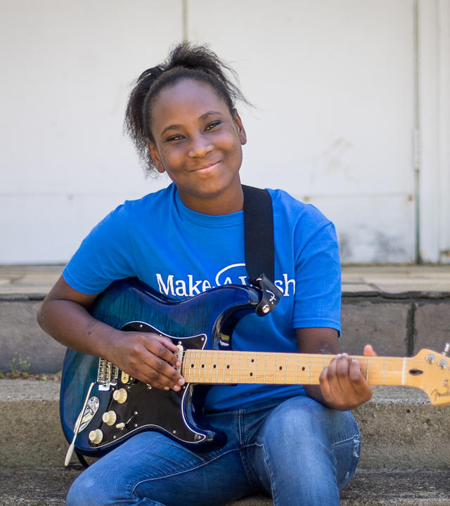 Wish kid Deonna holding a guitar wearing a Make-A-Wish t-shirt smiling